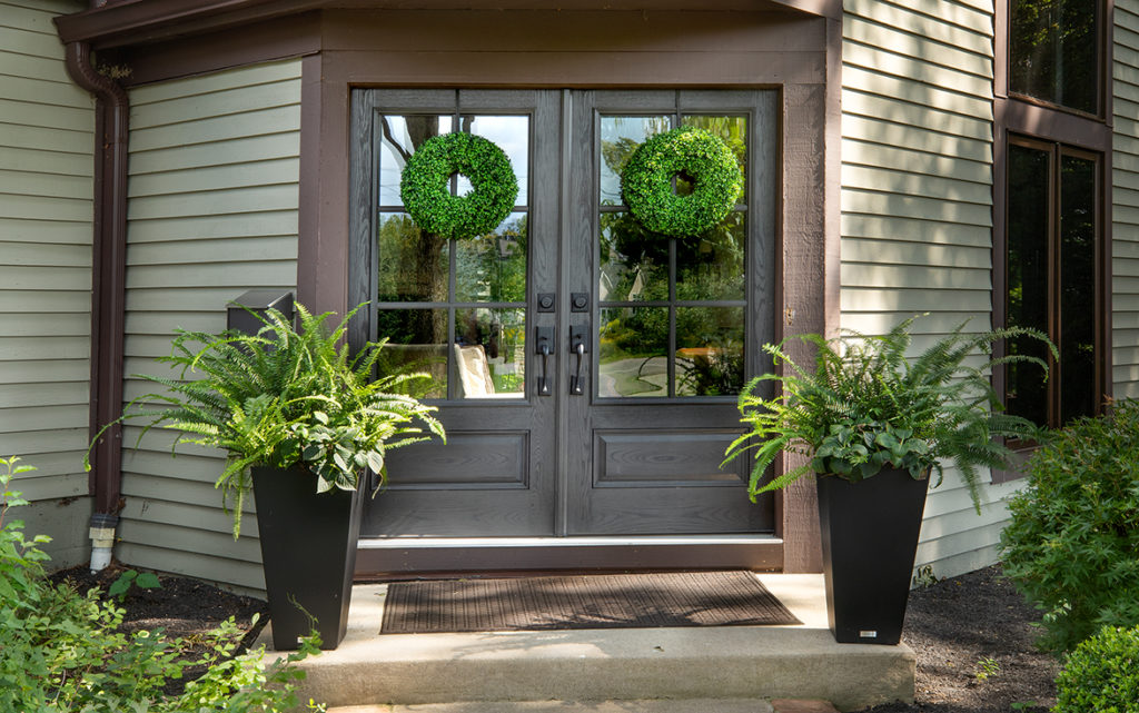 A double front door by Epic Group. The front door is dark gray with a wood texture and features two prominent windows on both doors. There are two green wreaths on both windows with two matching porch plants on either end of the doors.
