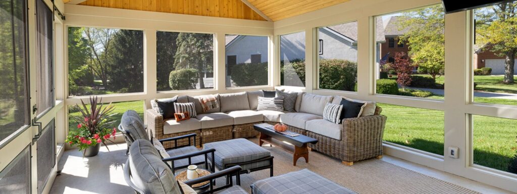 Screen porch addition with a large sectional couch, 2 outdoor friendly. chairs with matching ottomans. The wood paneled ceiling can be seen at the top of the image as well as the door leading to the patio.